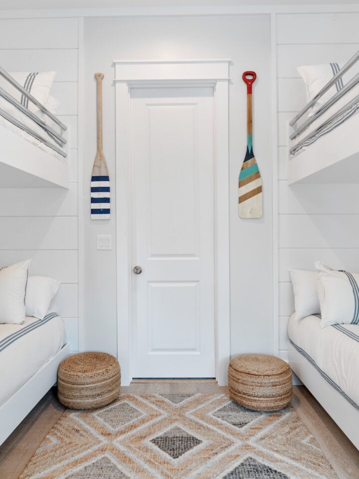 Bright, nautical-themed bedroom with white bunk beds and decorative paddles. Cozy and clean aesthetic, featuring patterned cushions and round woven stools.