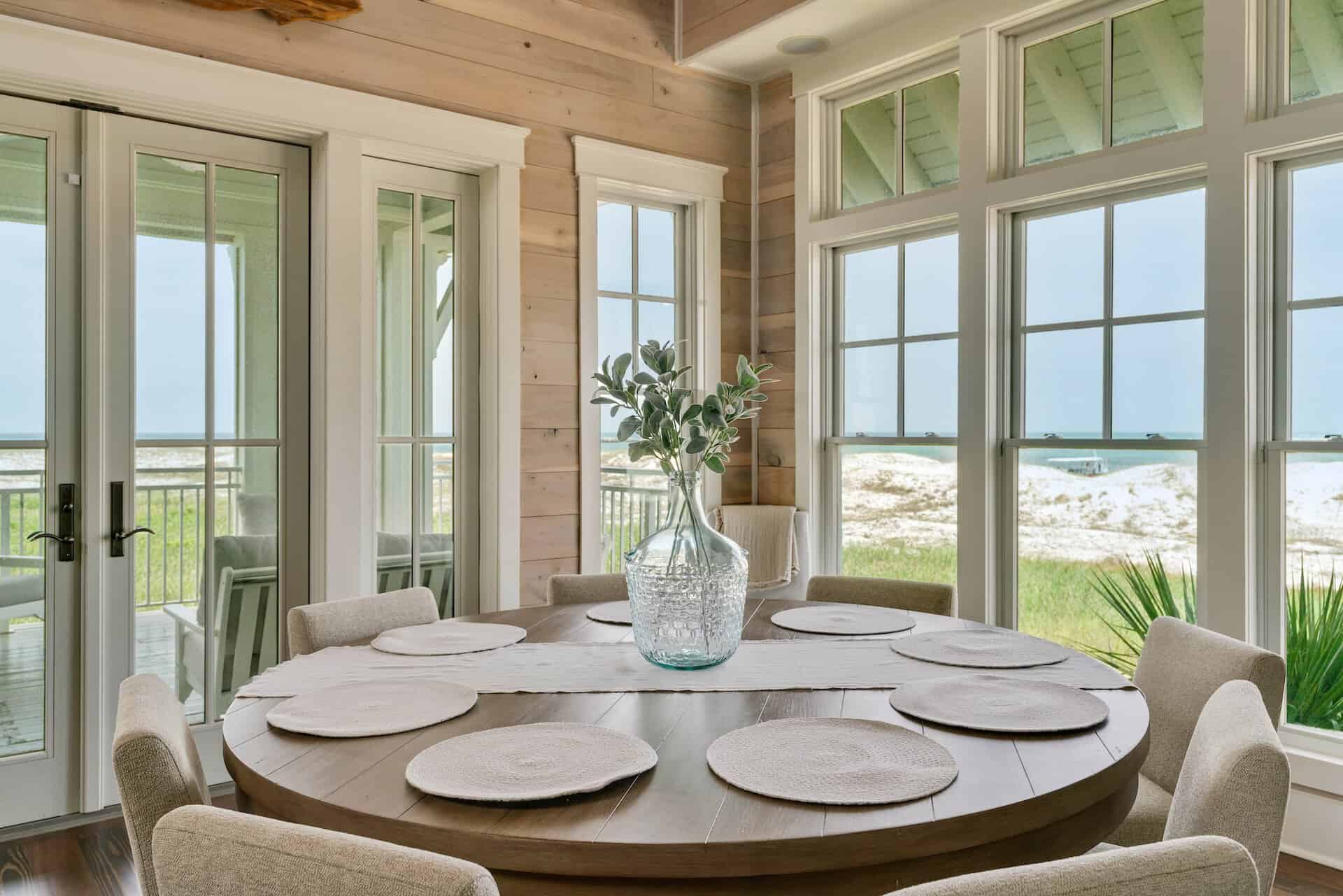 This residential interior design features a bright dining room with circular table set for six, large windows showing the sandy beaches of Santa Rosa Beach, FL by Kristin Midgett Design.