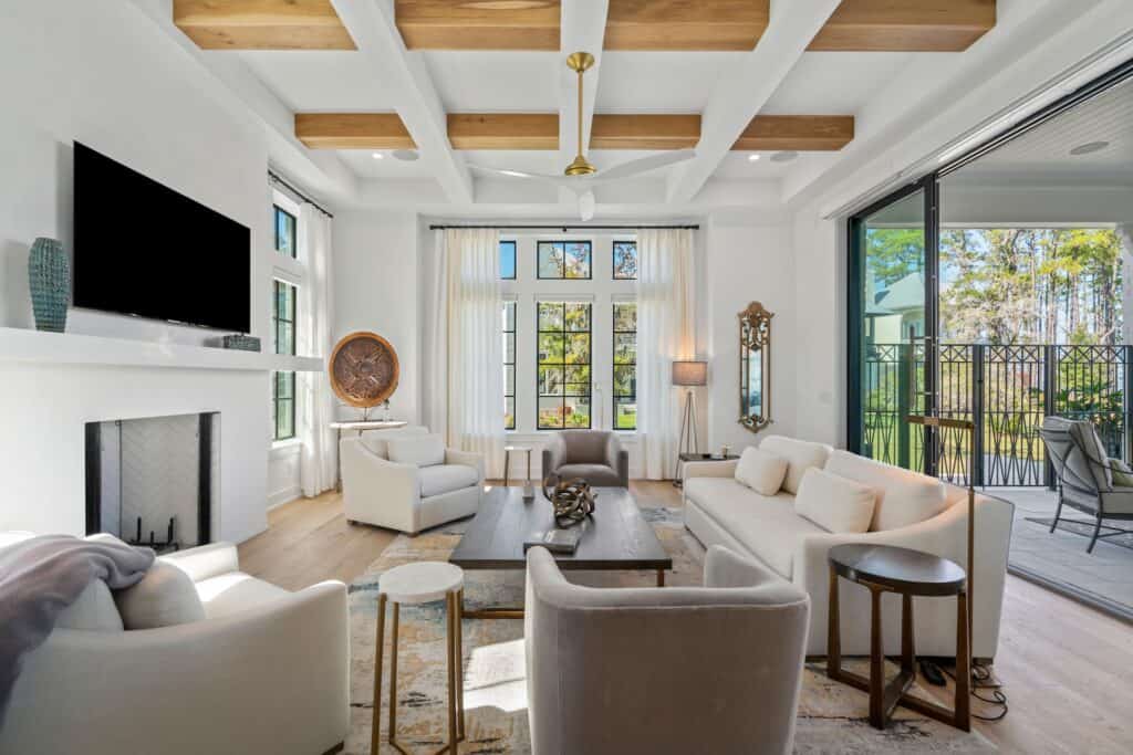 Modern living room featuring white furniture, wooden beams, large windows, and a fireplace. Sliding doors open to a balcony with garden views.