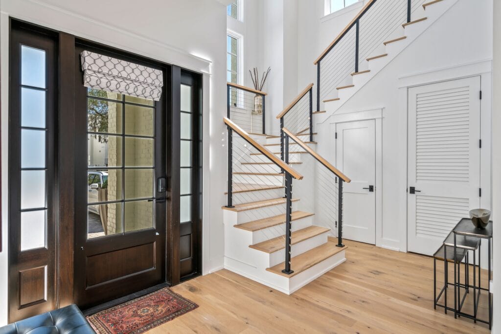 Modern entryway with wooden floors and staircase, featuring a black-framed glass door, patterned rug, and simple decorative accents on a side table.