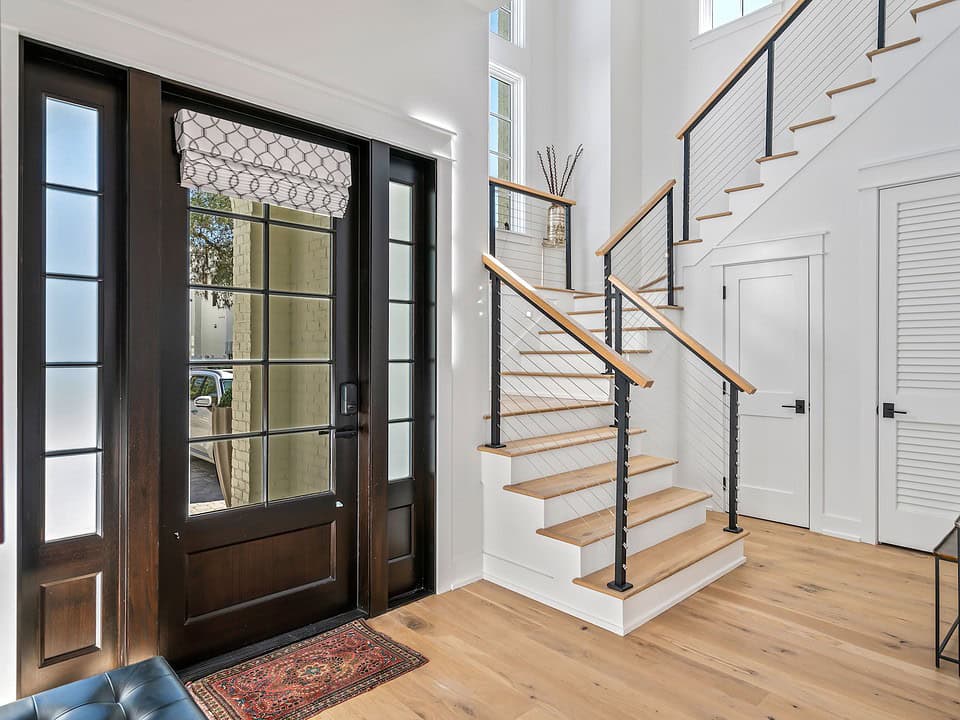 Modern entryway with wooden floors and staircase, featuring a black-framed glass door, patterned rug, and simple decorative accents on a side table.