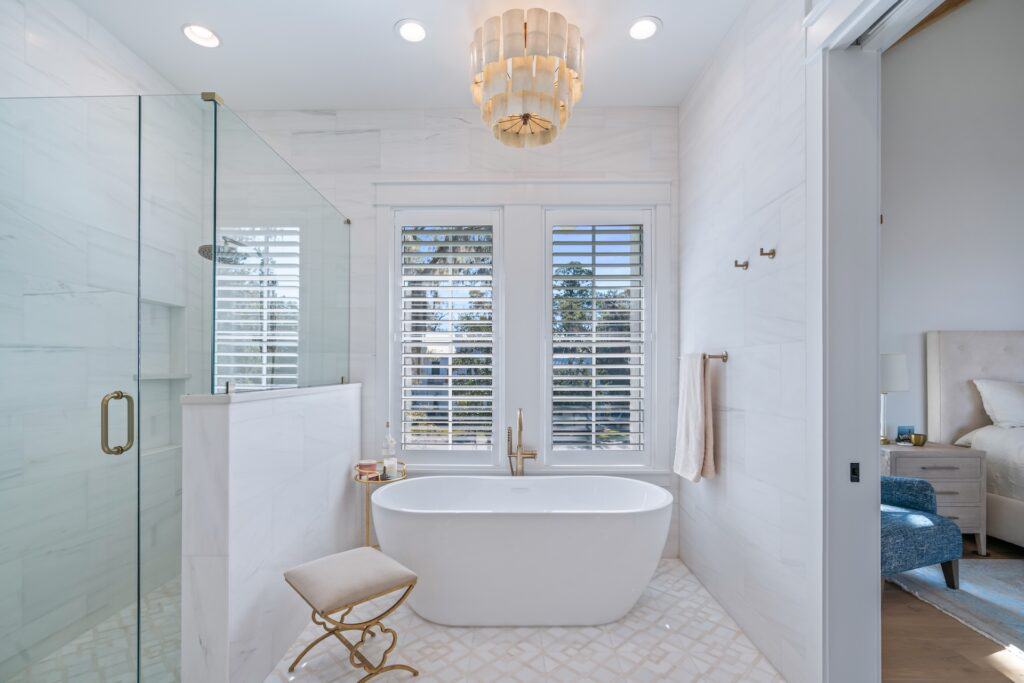 Elegant bathroom features a freestanding bathtub, glass shower, plush stool, and luxurious chandelier, with sunlight filtering through shuttered windows onto tiled floor.