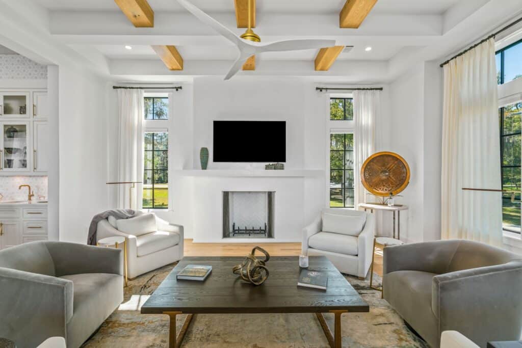 Modern living room with white walls, comfortable chairs, wooden beams, large windows, and a dark coffee table displaying decor items and books. Bright and inviting.