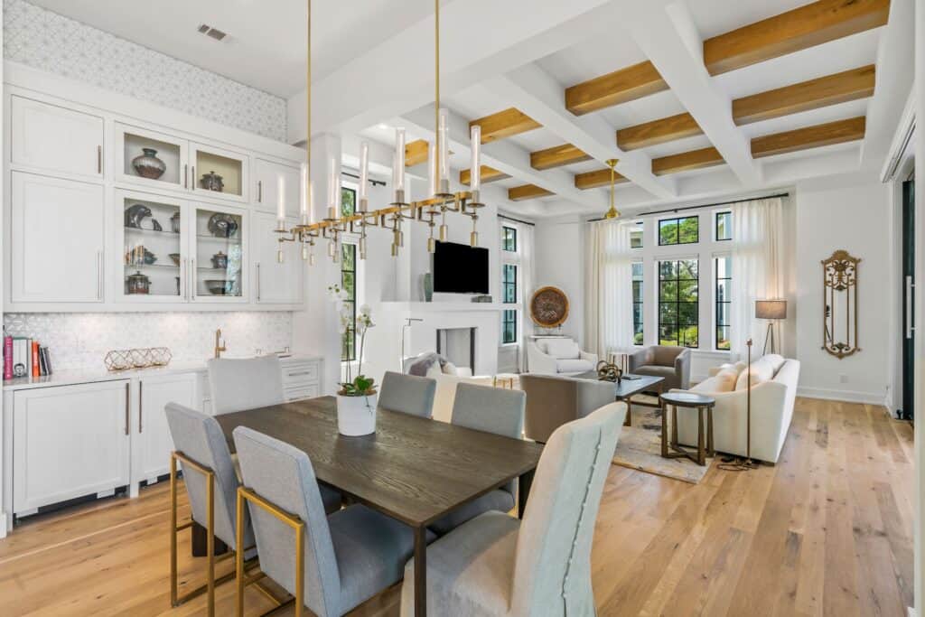 Modern living space featuring wooden beams, chic lighting, white cabinetry, and beige furnishings in a bright, open-concept layout with large windows.