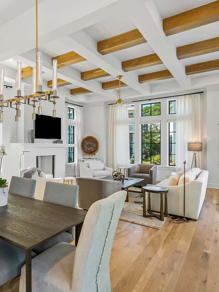 Modern living space featuring wooden beams, chic lighting, white cabinetry, and beige furnishings in a bright, open-concept layout with large windows.