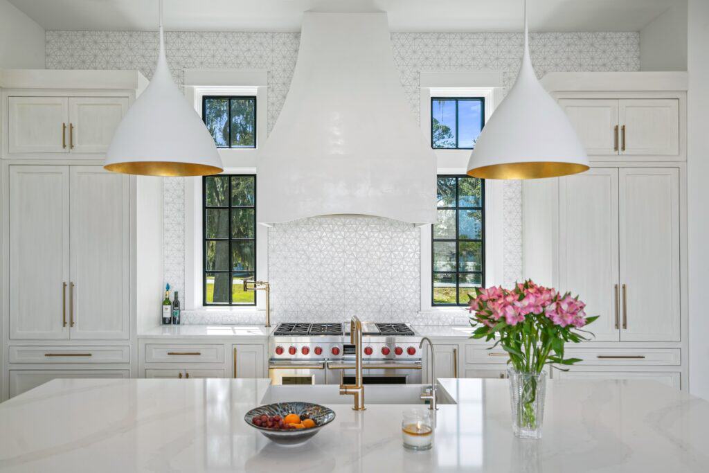 A modern kitchen in Churchill Oaks, Santa Rosa Beach, FL features white cabinetry, dual pendant lights, a central island with a vase of pink flowers, and a stainless steel stove.