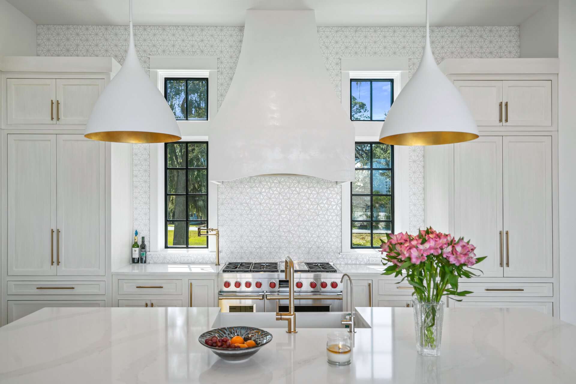 A modern kitchen in Churchill Oaks, Santa Rosa Beach, FL features white cabinetry, dual pendant lights, a central island with a vase of pink flowers, and a stainless steel stove.