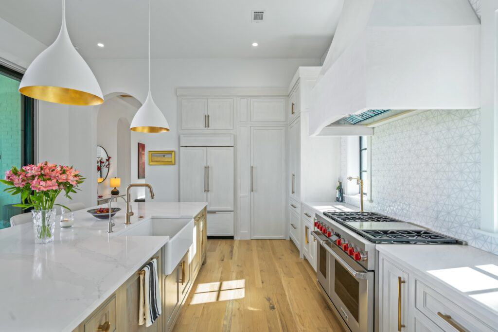 A modern kitchen with white cabinetry, large island, elegant lighting, and hardwood flooring. Flowers and decorative items add a touch of color.