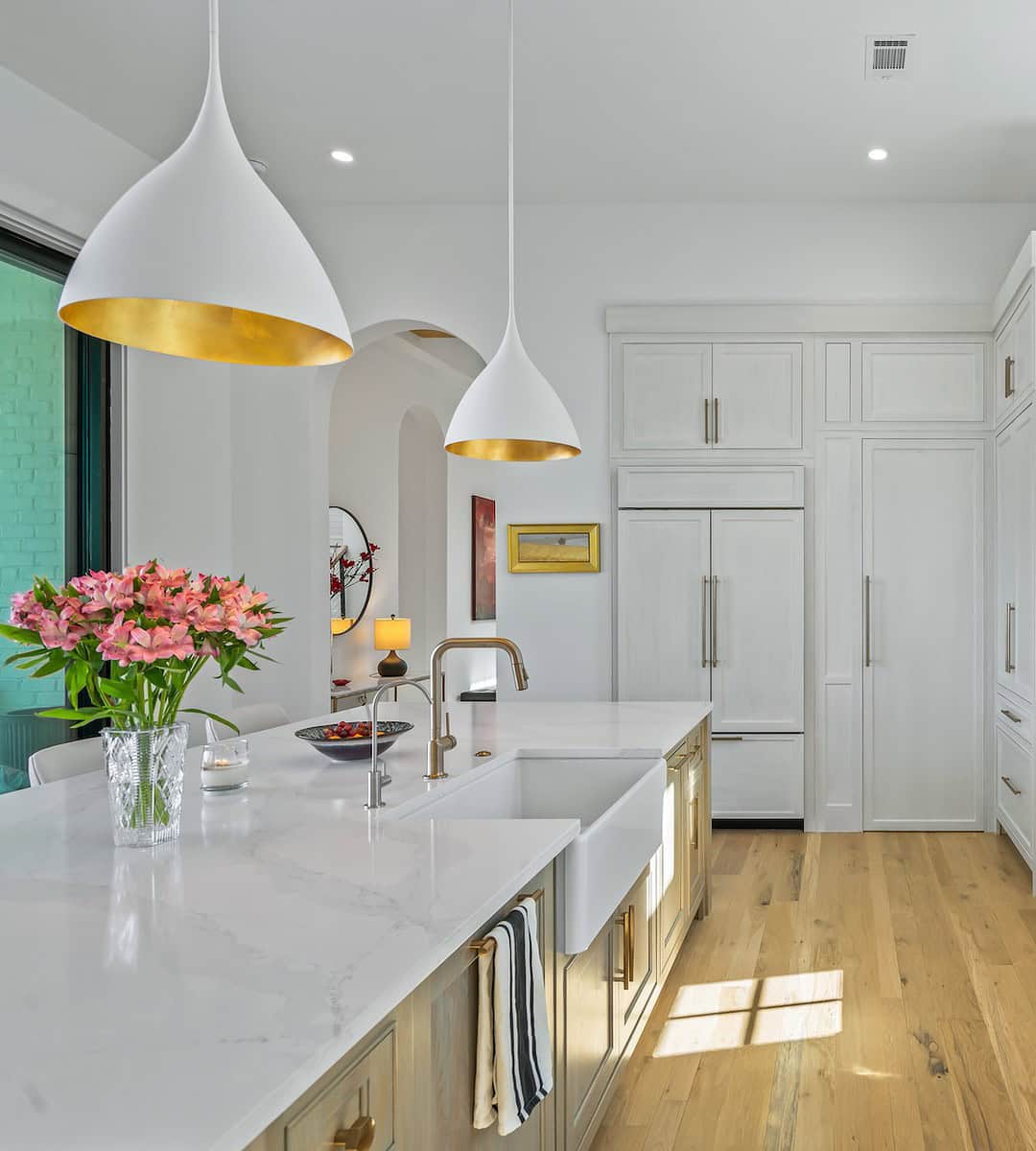 A modern kitchen with white cabinetry, large island, elegant lighting, and hardwood flooring. Flowers and decorative items add a touch of color.