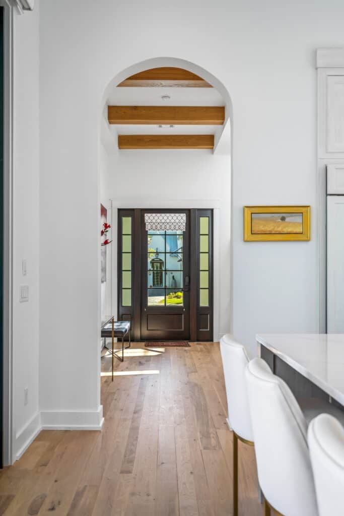 This modern interior hallway in Santa Rosa Beach, FL features wooden floors, an arched doorway, wall art, and a stylish glass-paneled front door with sunlight streaming in. Residential interior designer services provided by Kristin Midget Design.