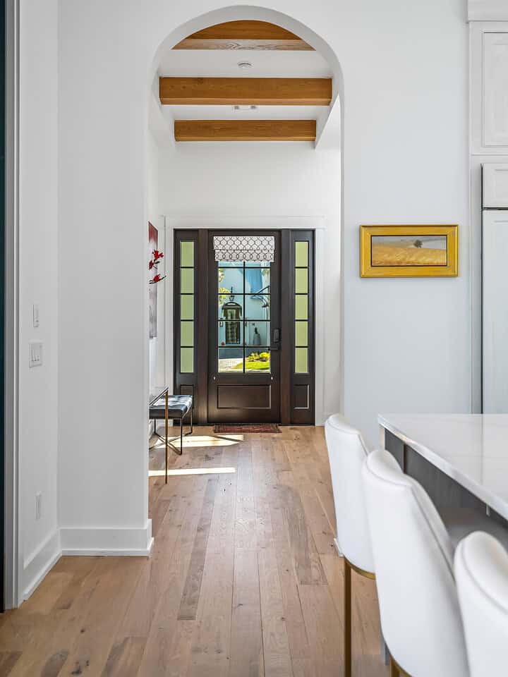 This modern interior hallway in Santa Rosa Beach, FL features wooden floors, an arched doorway, wall art, and a stylish glass-paneled front door with sunlight streaming in. Residential interior designer services provided by Kristin Midget Design.