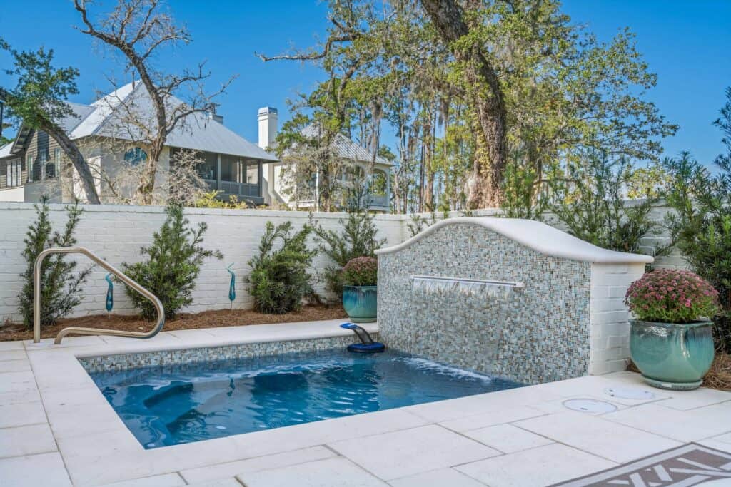 Small backyard pool with cascading waterfall feature, surrounded by trees, shrubs, and decorative pots, adjacent to a house with a tin roof.