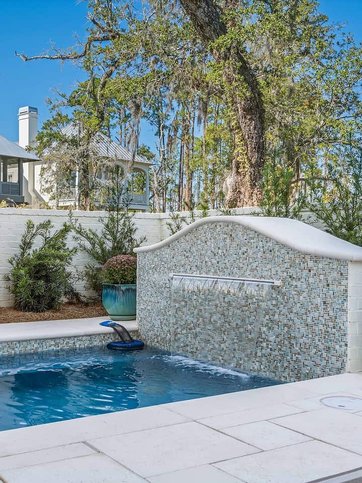 Small backyard pool with cascading waterfall feature, surrounded by trees, shrubs, and decorative pots, adjacent to a house with a tin roof.