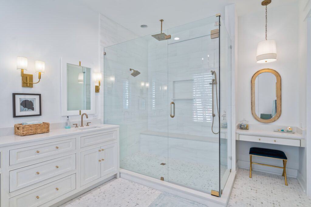 A modern bathroom features a glass-enclosed shower, white cabinetry, gold accents, and a small vanity area with an oval mirror and stool.