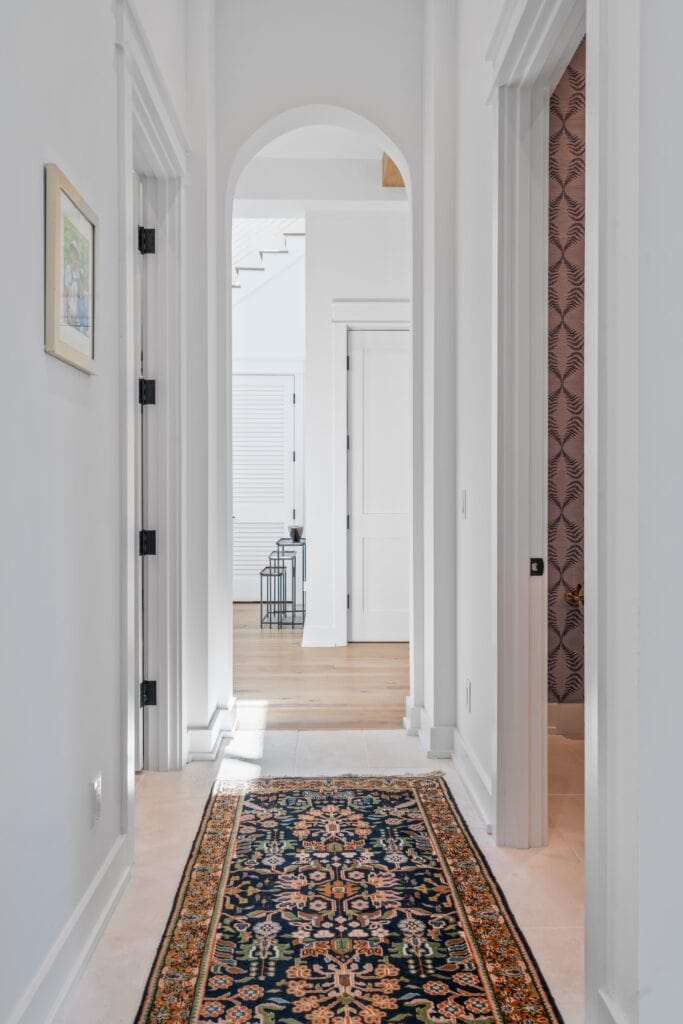 A hallway with a patterned rug, white walls, and doors. Natural light illuminates the wooden floor and decorative objects at the end.