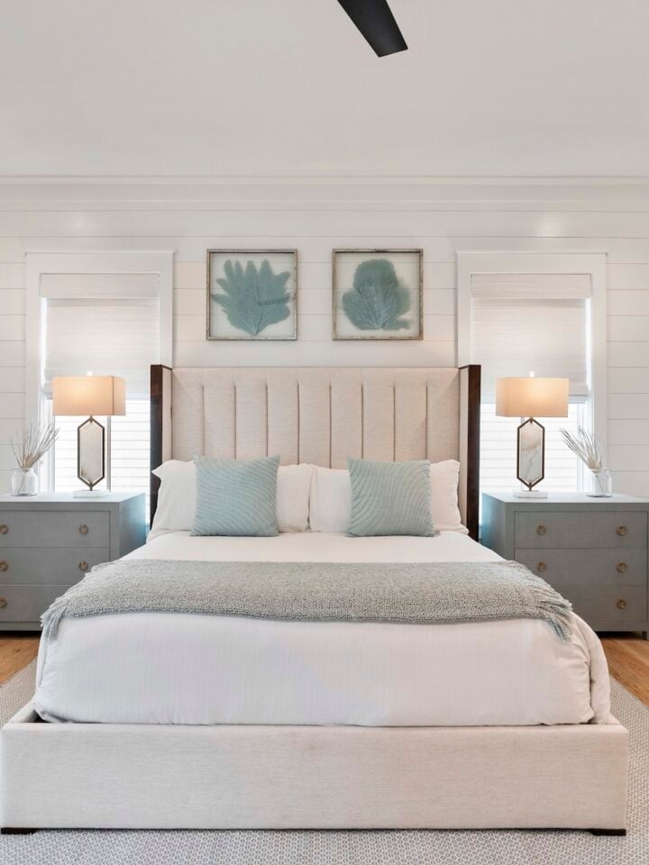 Bright bedroom with a beige bed, soft blue pillows, matching chairs, neutral decor, two lamps, and framed leaf prints on white walls.