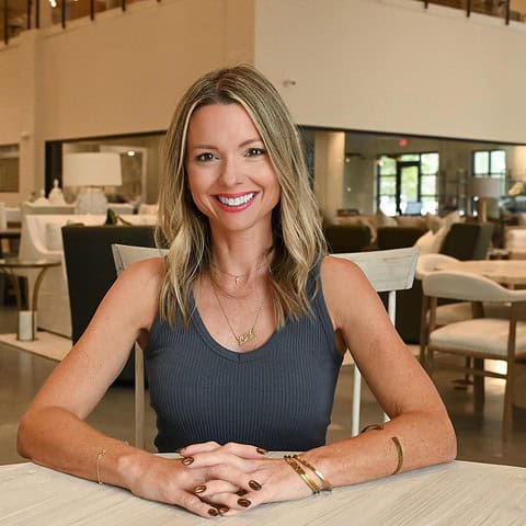 Kristin Midgett, Interior Designer in Santa Rosa Beach FL, sits at a table, smiling, in a modern, stylishly furnished interior space with chairs and decorative lighting.