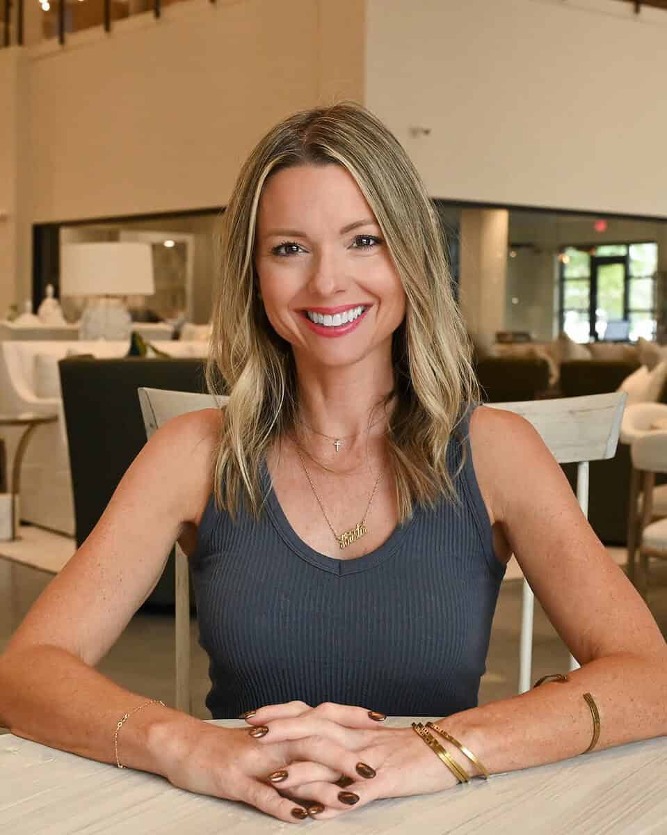 Kristin Midgett, Interior Designer in Santa Rosa Beach FL, sits at a table, smiling, in a modern, stylishly furnished interior space with chairs and decorative lighting.
