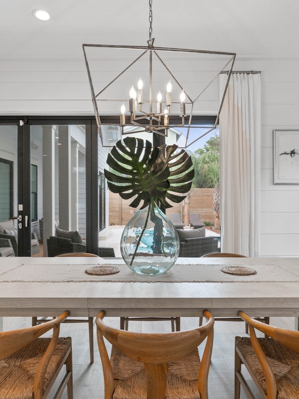 A modern dining area with wooden chairs, large glass vase centerpiece, and geometric chandelier, overlooking outdoor seating through sliding glass doors.
