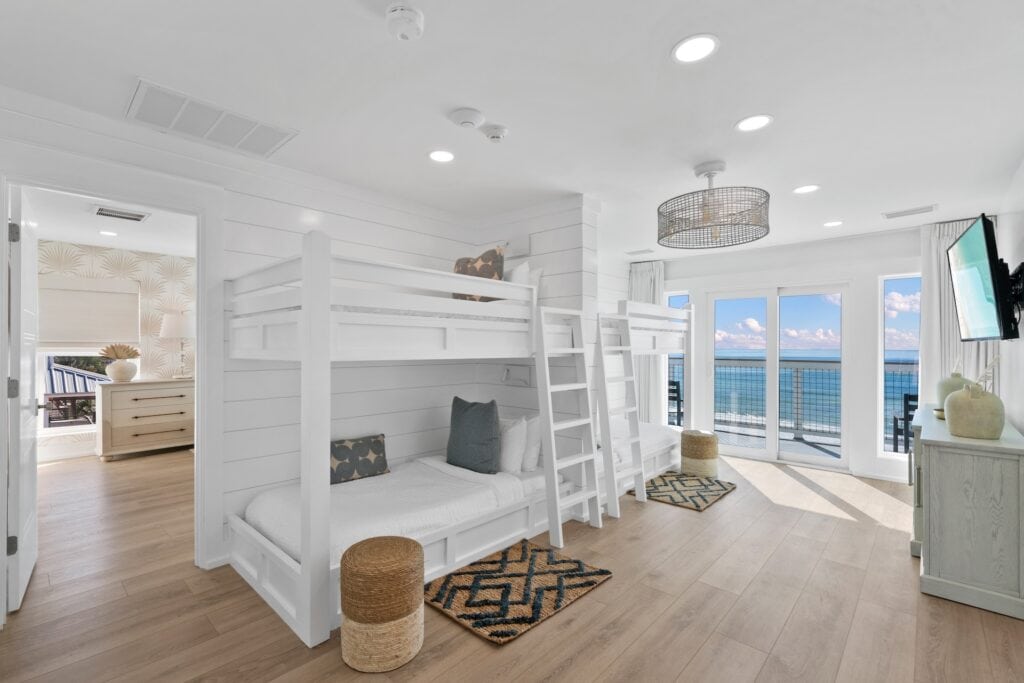 Bright coastal bedroom with white bunk beds, hardwood flooring, ocean view, and light-colored decor. Sliding doors open to a sunny balcony.