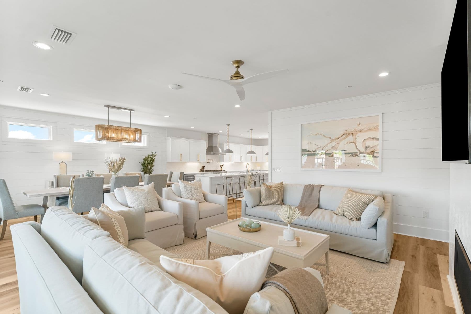 Spacious modern living room with light neutral tones, comfortable seating, dining area, and open kitchen. Decorative art piece and ample natural light.