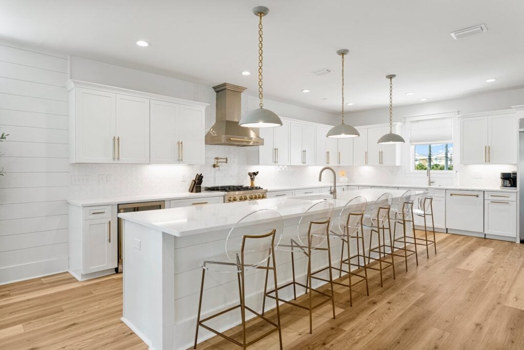 Modern kitchen with white cabinets, pendant lighting, stainless steel appliances, and a long island with transparent bar stools on wooden flooring.