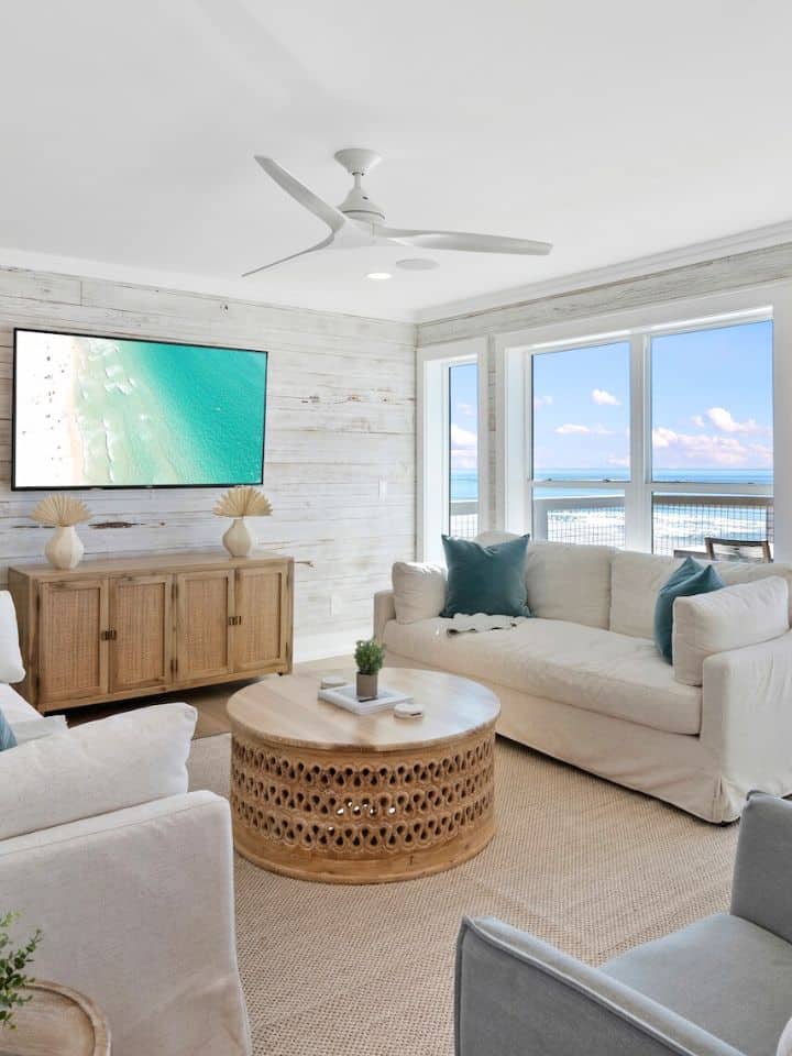 Bright living room with beach-themed decor, featuring a large window with ocean view, a TV, ceiling fan, cozy sofas, and wooden furniture.