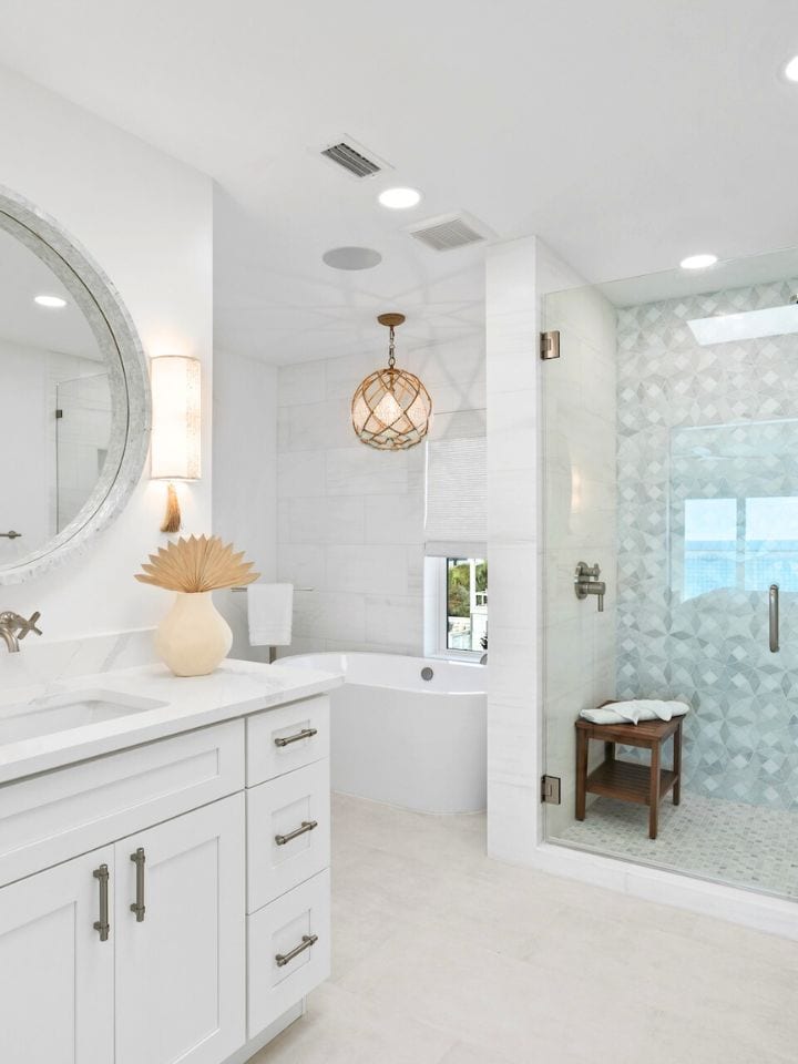 Modern bathroom with a round mirror, pendant light, large bathtub, and glass shower. Bright, minimalistic design with white cabinetry and tiled floor.
