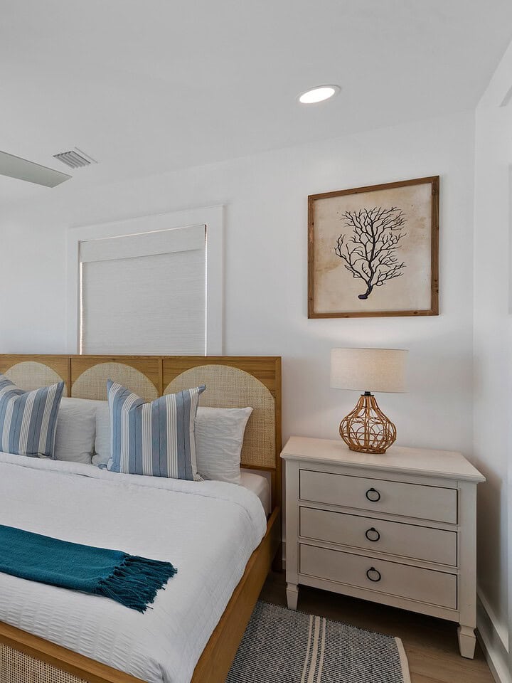 A bright, airy bedroom with ocean views, decorated with coastal art, featuring a large bed, ceiling fan, and windows overlooking the beach.