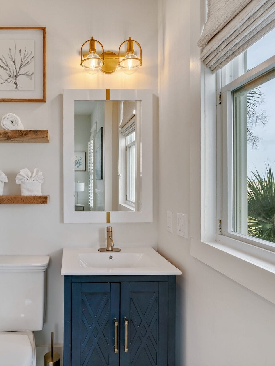 Modern bathroom with glass shower, white sink, and navy cabinet. Shelves hold towels. Window view shows greenery. Bright, clean aesthetic.