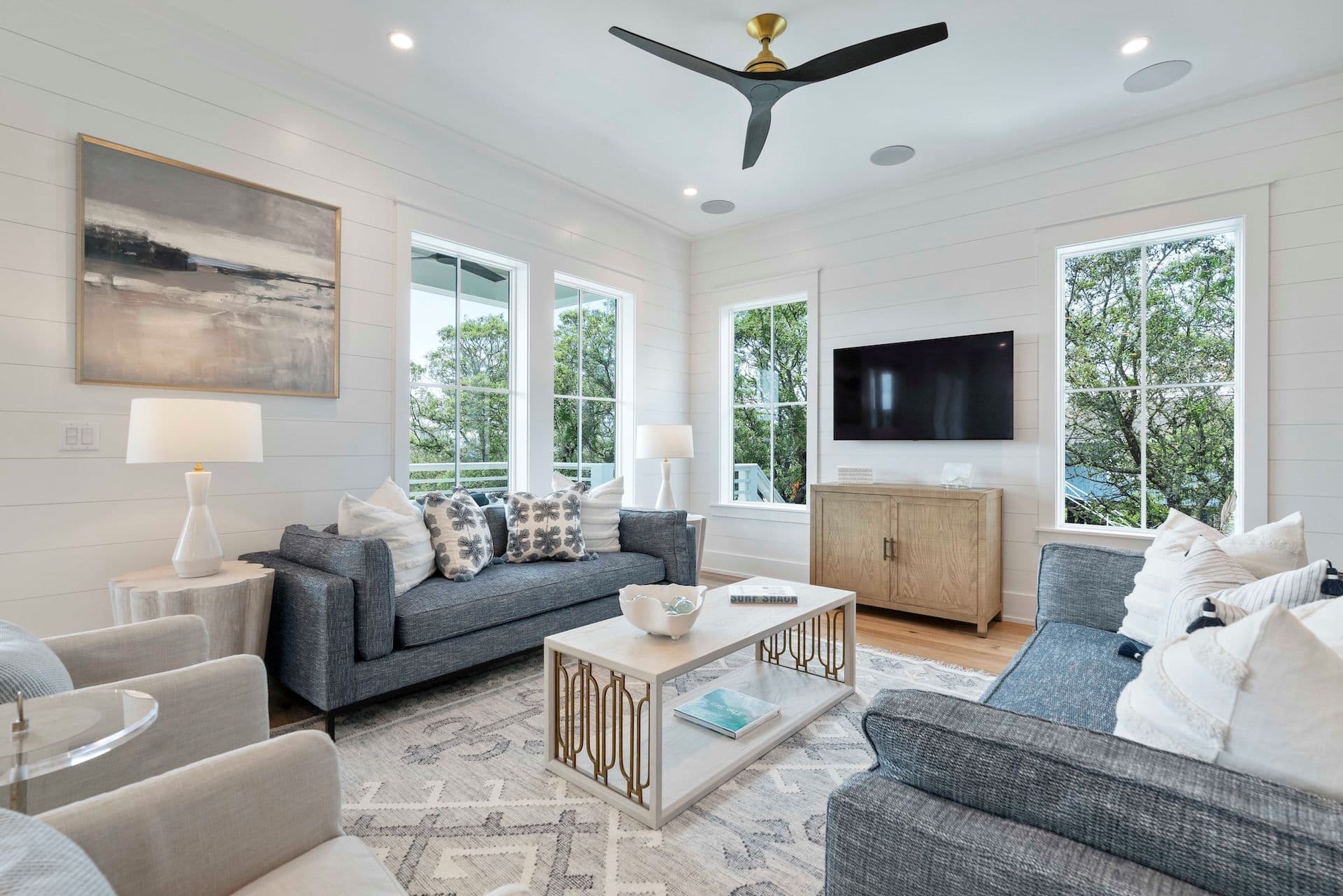 A modern living room features two sofas, a coffee table, large windows, wall art, a TV, and a ceiling fan.
