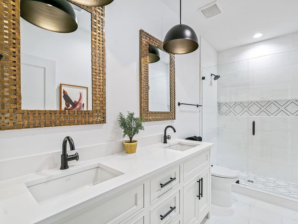 Modern bathroom features twin sinks, woven framed mirrors, black faucets, pendant lights, glass shower enclosure, geometric tile design, and a potted plant.
