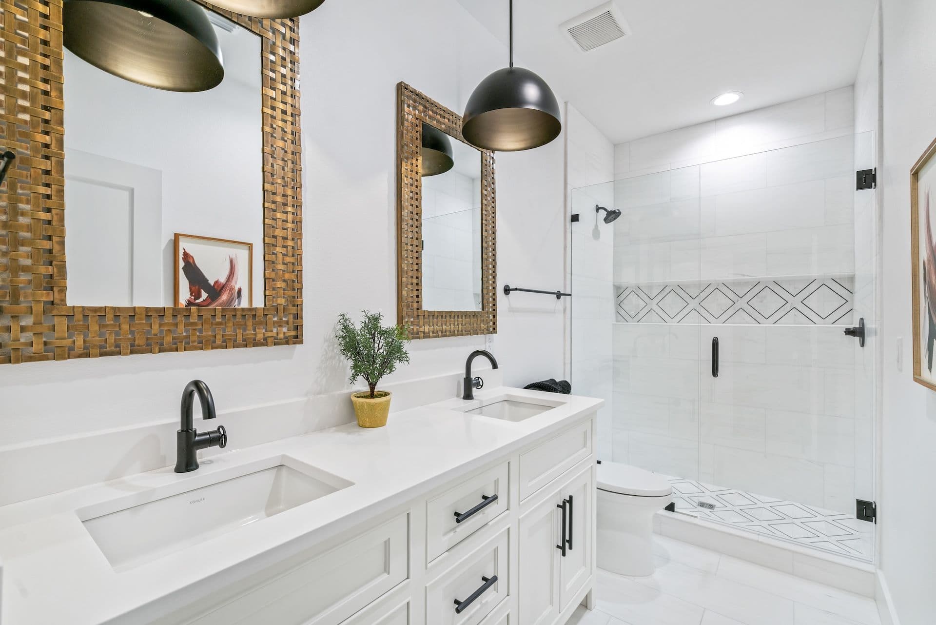 Modern bathroom features twin sinks, woven framed mirrors, black faucets, pendant lights, glass shower enclosure, geometric tile design, and a potted plant.