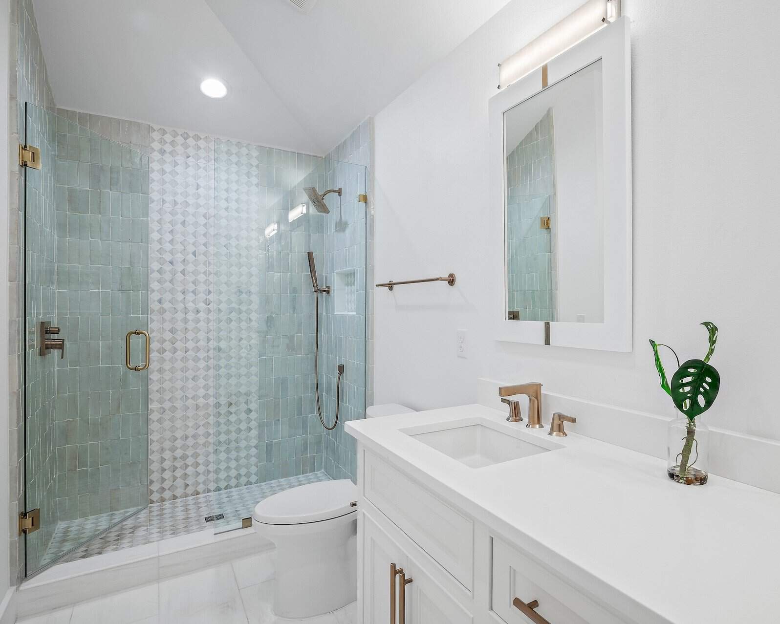 Modern bathroom with glass shower, white vanity, large mirror, and gold fixtures. Decorative plant in a glass jar on the countertop.