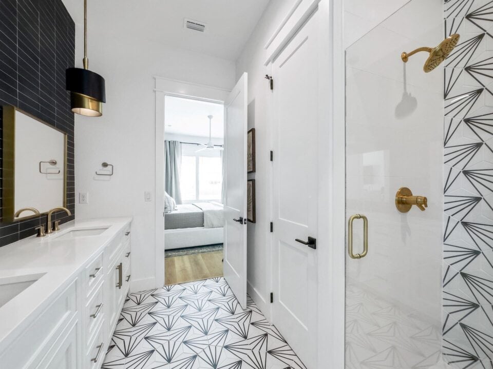 Modern bathroom with patterned tile, white cabinetry, gold fixtures, and pendant lighting. Open door reveals a bedroom with large windows and curtains.