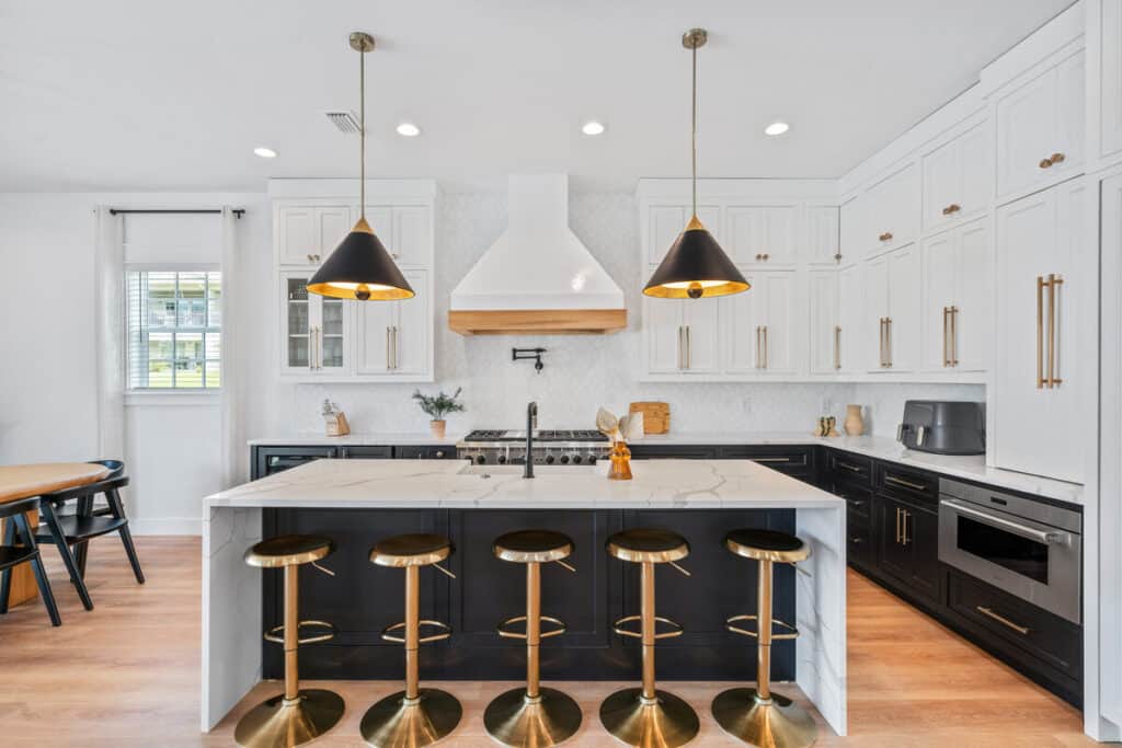 Modern kitchen with black and white cabinetry, brass fixtures, marble countertop, bar stools, pendant lights, and wooden flooring. Oven and air fryer visible.