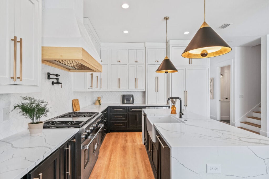 Modern kitchen featuring white cabinets, black base units, marble countertops, pendant lights, stove, and sink. Wooden flooring and a staircase visible.