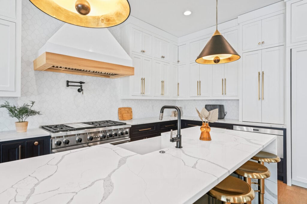 Modern kitchen with marble countertops, gold pendant lights, stainless steel stove, and white cabinets. Stylish, elegant design with black and wood accents.