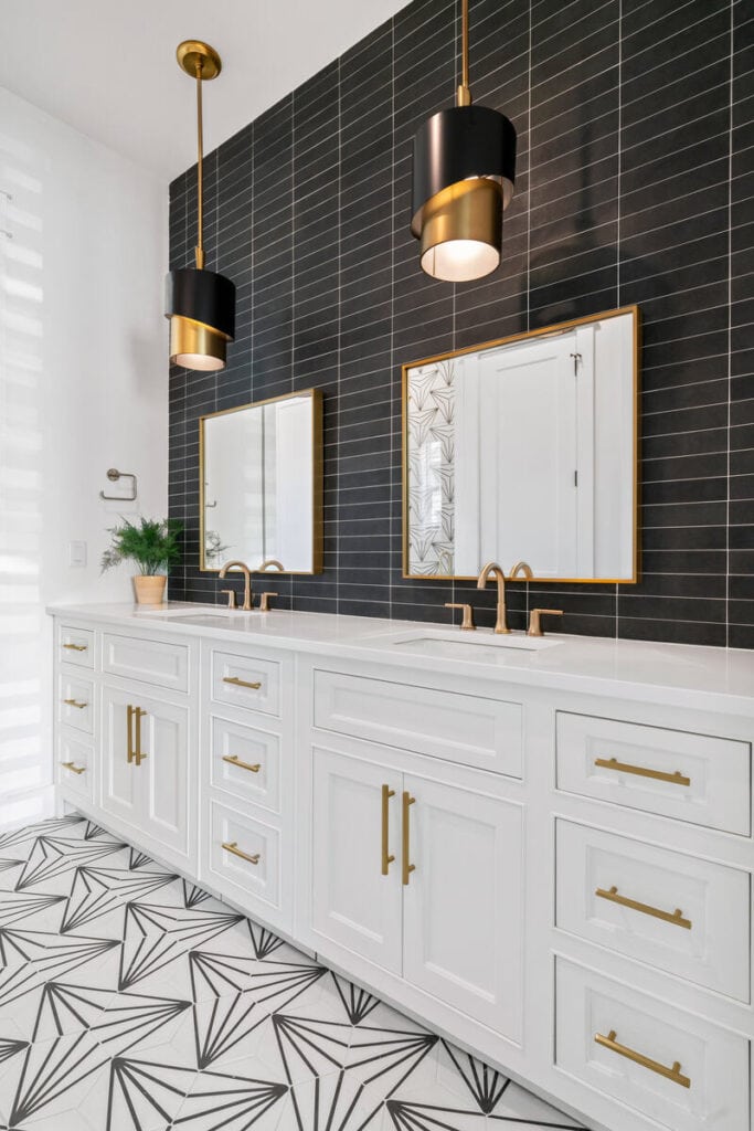 Modern bathroom with geometric floor tiles, dual white vanities, gold fixtures, black tile wall, large mirrors, and two stylish black pendant lights.