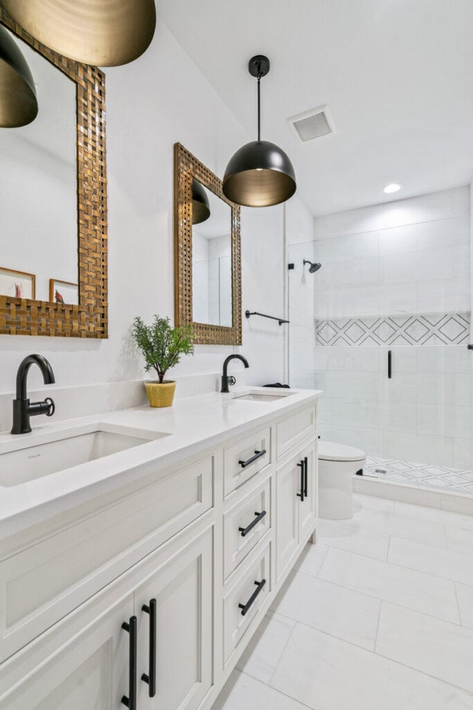 Modern bathroom with double sinks, brass mirrors, black fixtures, and glass shower. Features sleek white cabinetry and geometric tile accents, creating an elegant aesthetic.