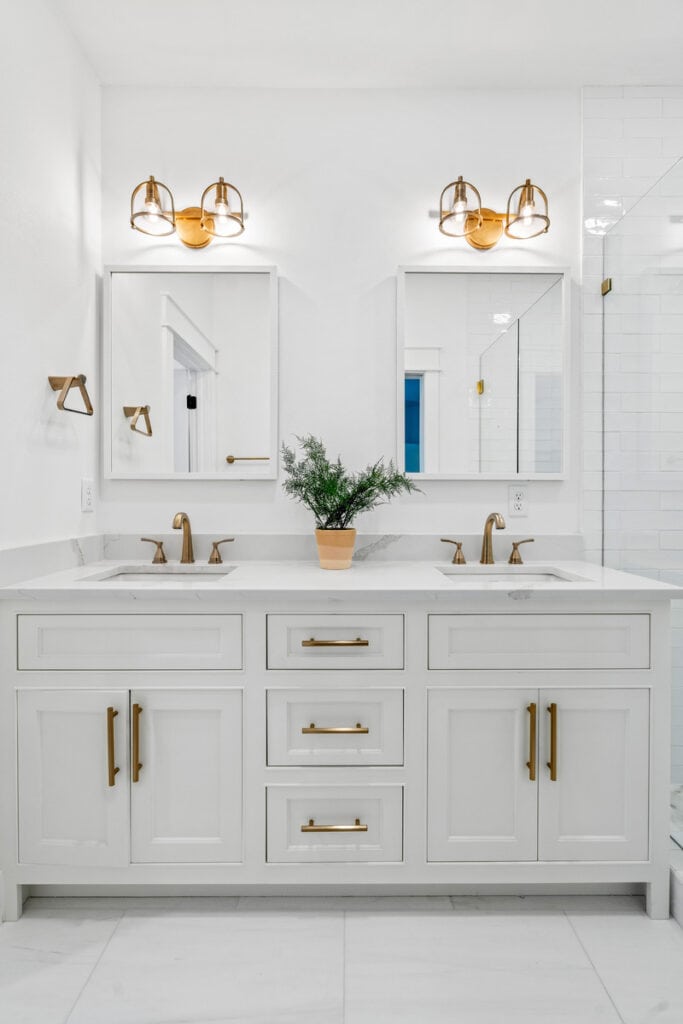 White bathroom with dual sinks, gold fixtures, mirrors, and a small potted plant on a marble countertop. Bright lighting enhances the modern design.