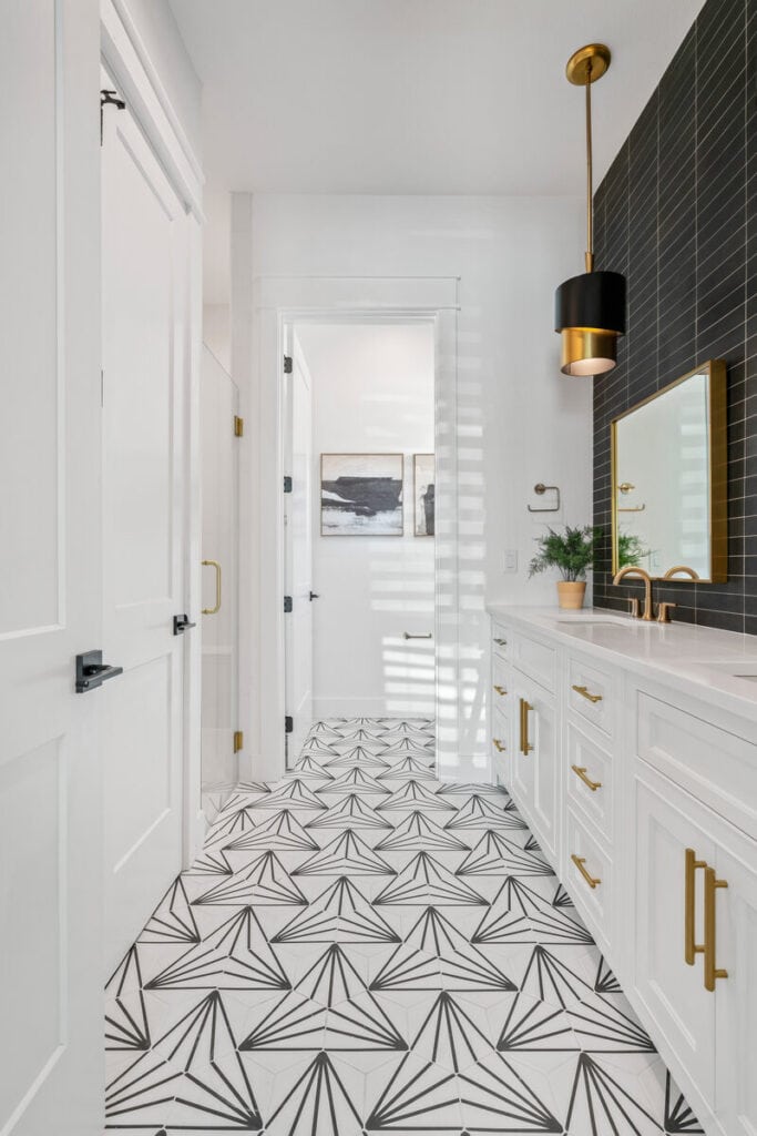 Modern bathroom with geometric floor tiles, white cabinetry, gold accents, black backsplash, and wall art. Natural light filters through windows, enhancing space.