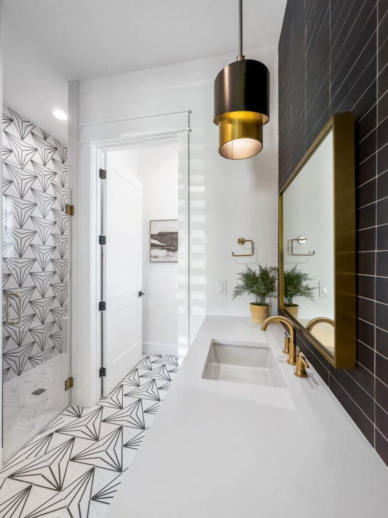 Modern bathroom with geometric tile patterns, a hanging pendant light, gold fixtures, a black-tiled mirror, and a white vanity.