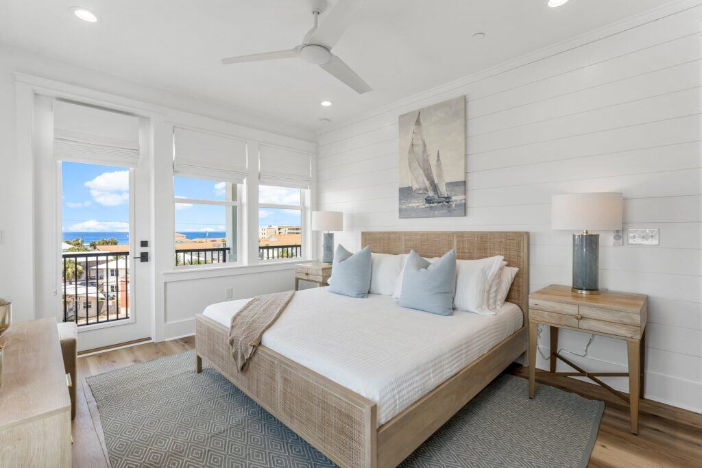 Bright bedroom with ocean view, featuring a wooden bed, nautical artwork, and light décor. Large windows offer abundant natural light and scenic vistas.