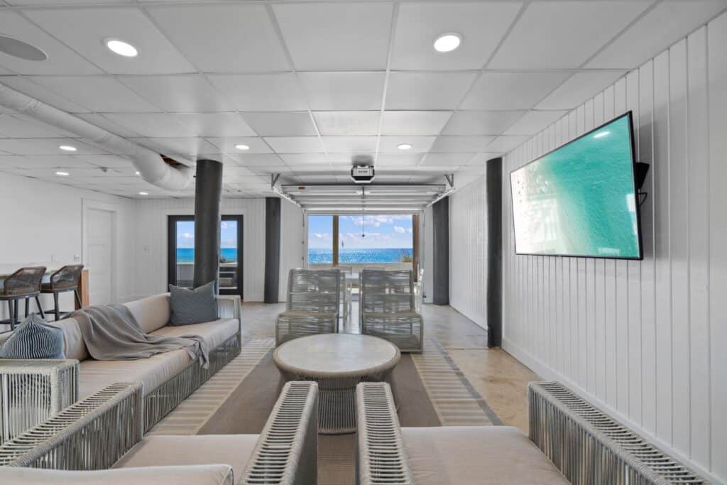 Modern beachfront living room with sleek furniture, large window view of the ocean, ceiling lights, and a wall-mounted television displaying a beach scene.