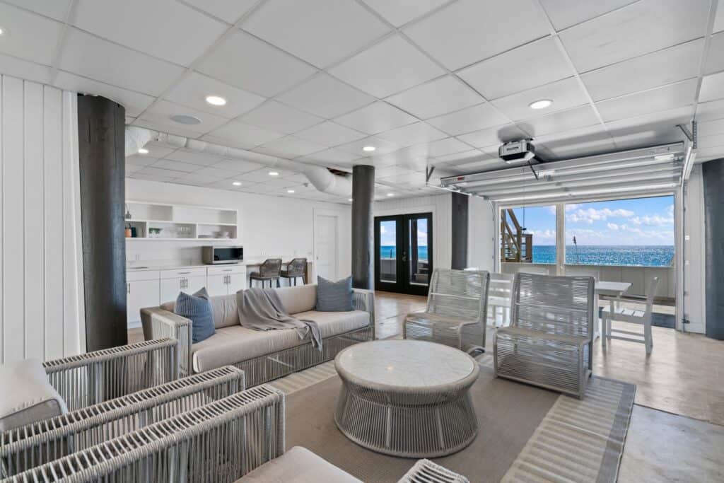Contemporary living space with ocean view features stylish furniture, wicker chairs, and open garage door, blending indoor comforts with outdoor scenery.