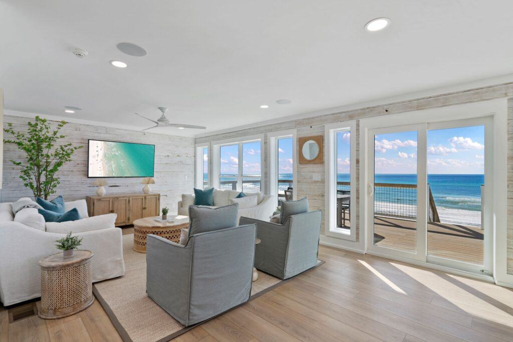 A bright living room with coastal decor, large windows offering ocean views, light wood floors, gray furniture, and a wall-mounted television.