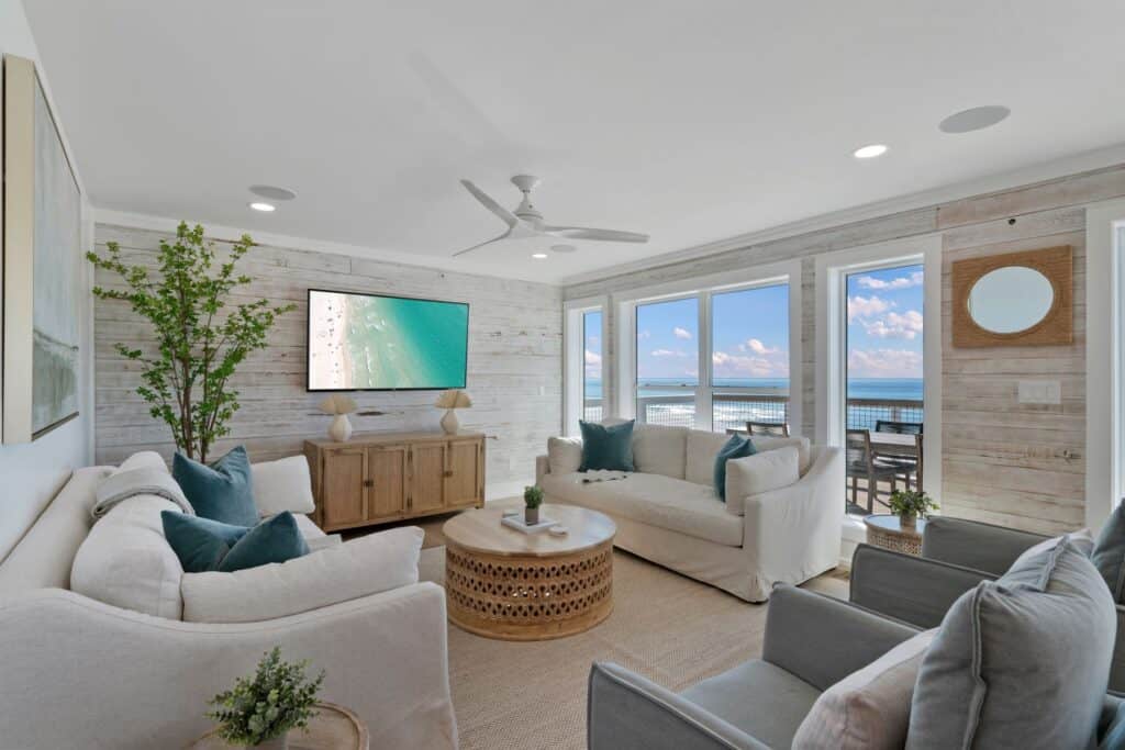Cozy living room with ocean view, neutral decor, white sofas, teal pillows, a wooden table, and a television showing a beach scene.