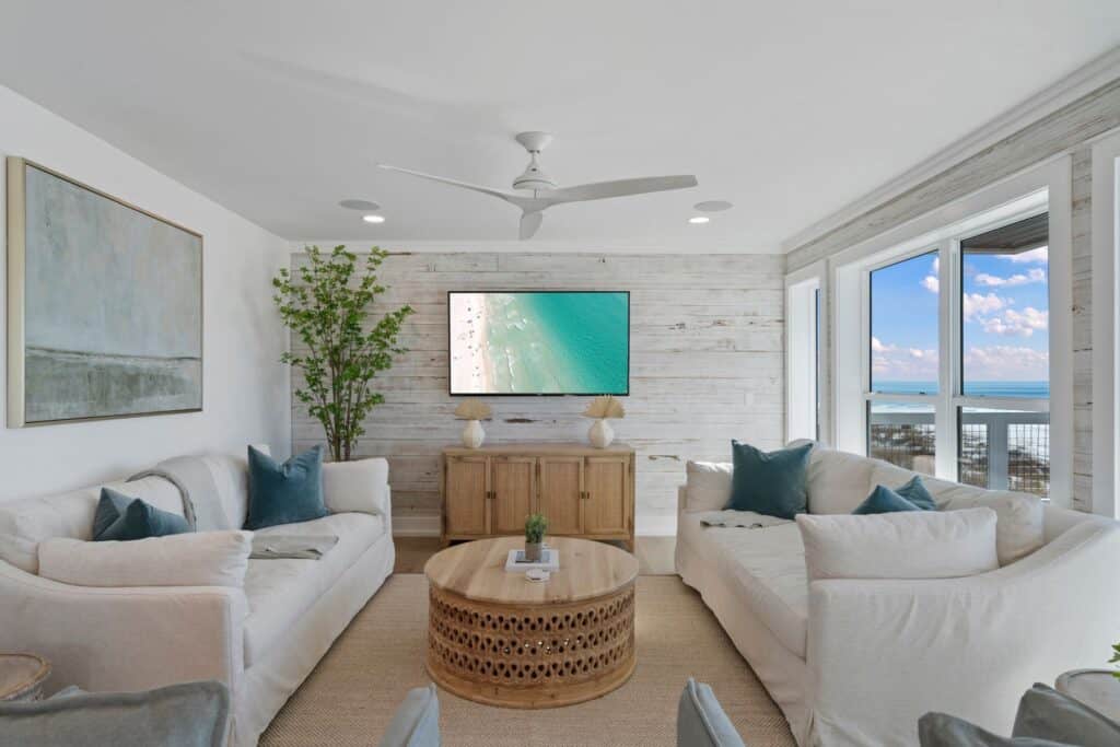 A cozy, modern living room features light sofas, a wooden coffee table, beach artwork, and large windows overlooking the ocean under a cloudy sky.