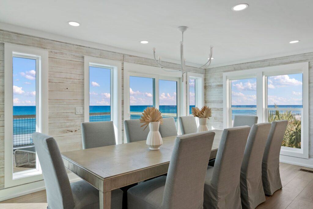 Spacious dining room with large windows, offering a stunning ocean view. Elegant decor includes a long table, high-backed chairs, and decorative vases.